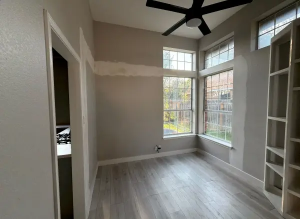Newly renovated bedroom with light gray vinyl plank flooring, fresh drywall and paint, built-in shelving, black ceiling fan, and large windows overlooking residential area in Austin, TX.