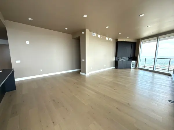 Finished high-rise apartment living area with new light wood flooring, freshly painted tan and cream walls, recessed lighting, and floor-to-ceiling windows overlooking cityscape in Austin, TX.