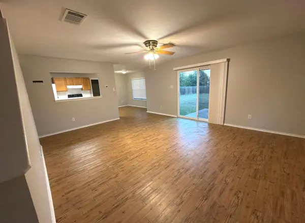 Clean living room with newly installed laminate or wood-look flooring, neutral walls, ceiling fan, kitchen visible through pass-through window, and glass sliding door to backyard in Leander, TX.