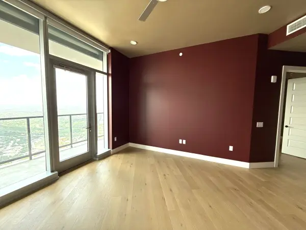 Finished high-rise apartment living room with deep burgundy accent wall, light wood flooring, recessed lighting, and floor-to-ceiling windows overlooking a distant cityscape in Austin, TX.