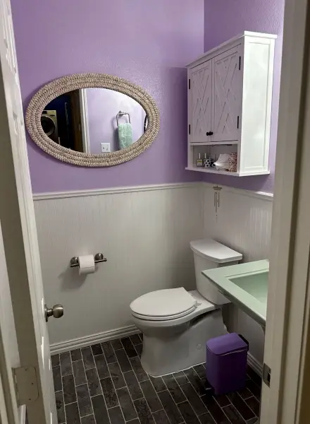 Completed bathroom renovation showing purple upper walls, white wainscoting, new oval mirror, white cabinetry, white toilet, light green sink vanity, and dark floor tile in Leander, TX.